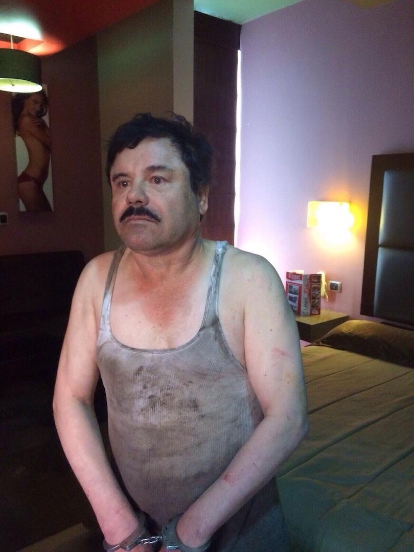 Ο Joaquín El Chapo Guzmán στέκεται δεμένος με χειροπέδες στην άκρη ενός κρεβατιού, στο μοτέλ Doux, μετά τη σύλληψή του. Το φανελάκι του είναι βρώμικο, λόγω της μετακίνησής του μέσω του αποχετευτικού συστήματος της πόλης.